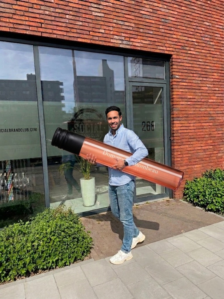 Lachende man houdt een reusachtige Domaine de Rombeau-wijntube vast voor het kantoor van The Social Brand Club.