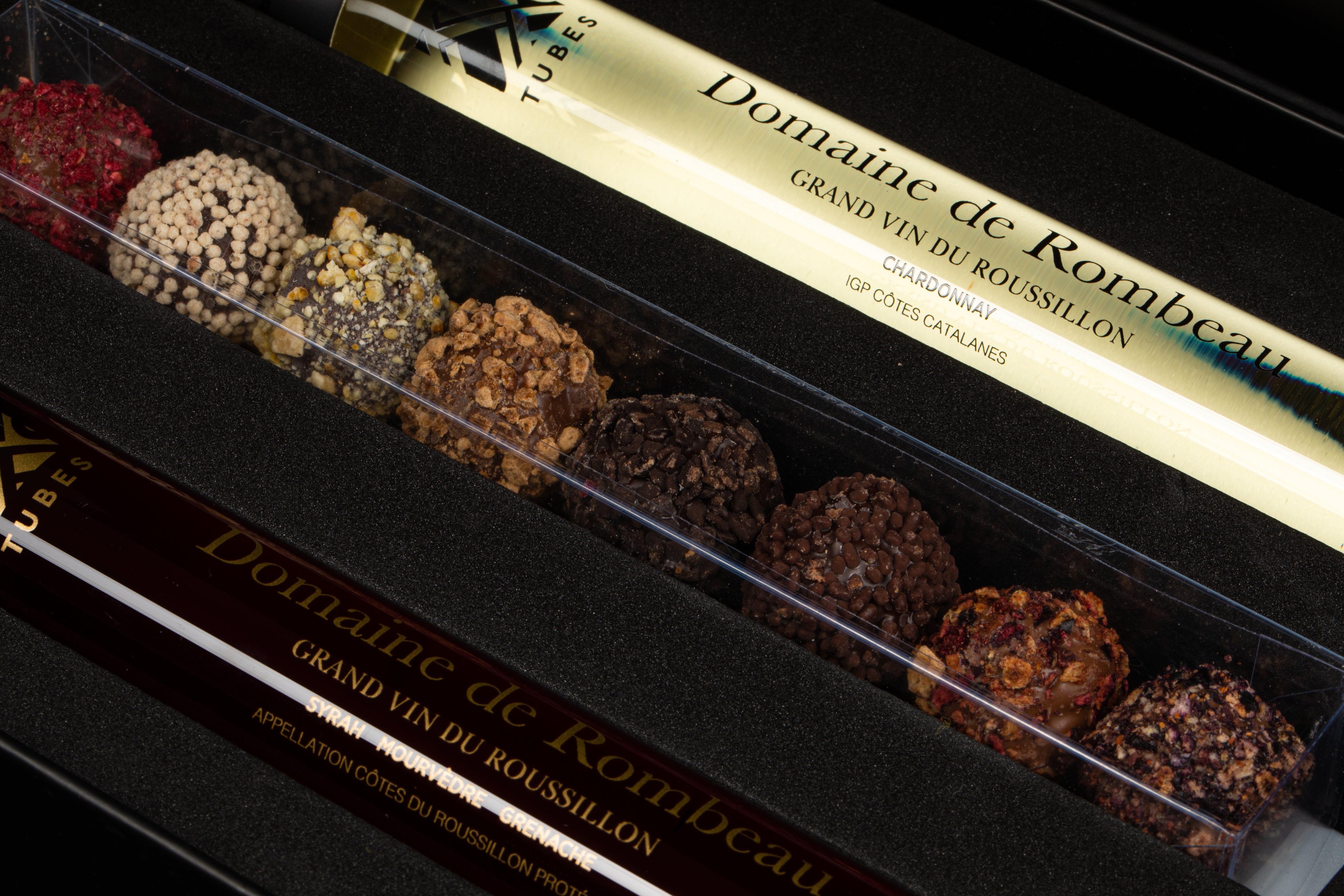 Rij gevarieerde chocoladetruffels met diverse toppings naast Domaine de Rombeau Chardonnay- en Syrah-wijntubes in een geschenkbox.