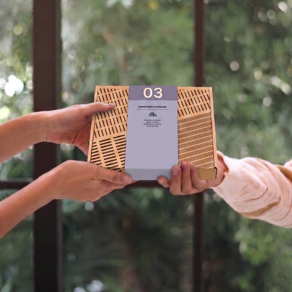Twee handen geven een kraftkartonnen kruidenbox met paarse band "Something's Cooking" door, voor een raam met groen erachter. 