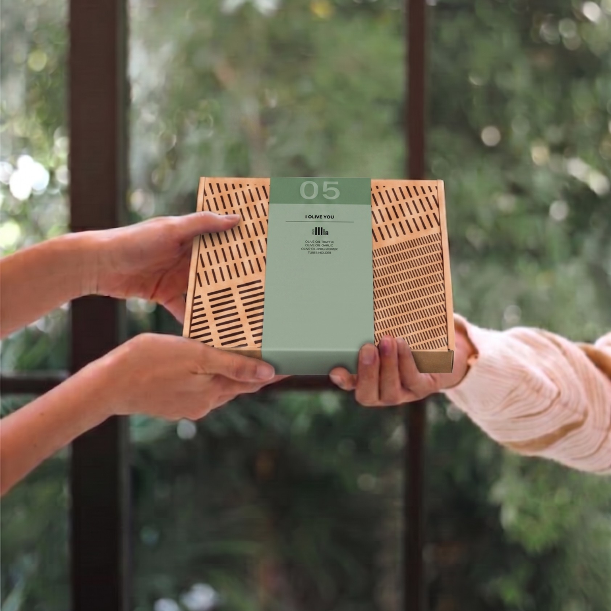 Alt-tekst: Twee handen geven een kraftkartonnen olijfolie-cadeaubox met groene band "I Olive You" door, voor een raam met groen. 