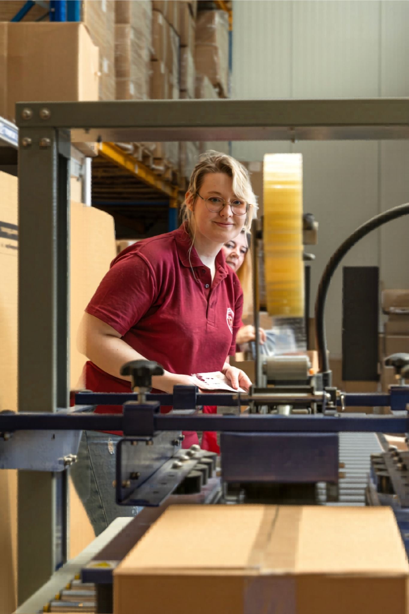 Vrouw met bril in bordeauxrode polo glimlacht bij een inpakmachine in een magazijn vol kartonnen dozen. 