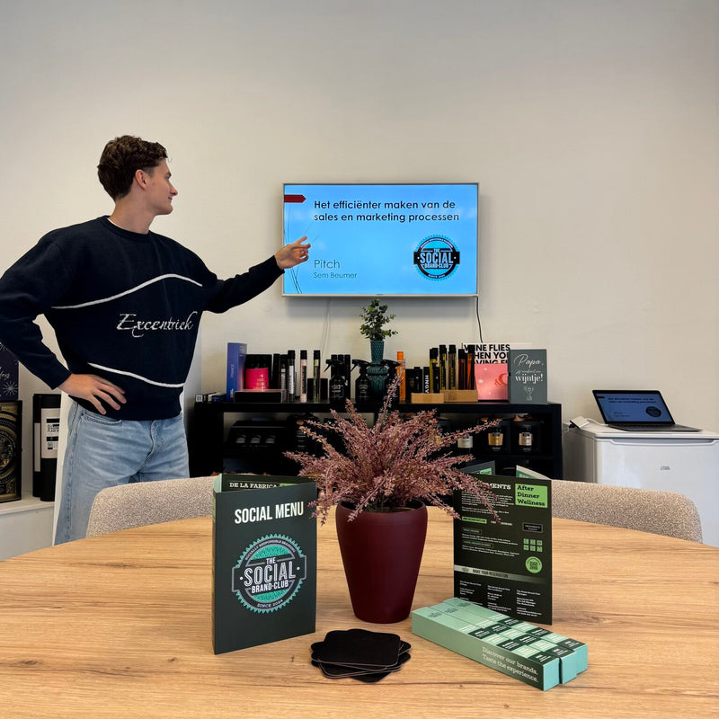 Jonge man wijst naar een scherm met een pitch van The Social Brand Club, met op de vergadertafel het Social Menu en merkproducten. 
